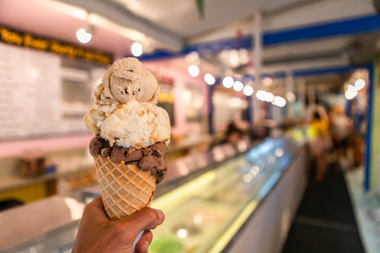 A close up and first person perspective of a man holding a waffle cone with scoops of ice cream at a shop, with blurry counter in the background