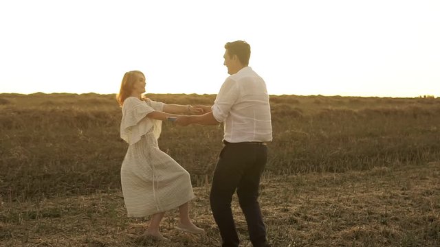 Slow motion of romantic couple holding hands and spin around. Woman in dress and man in suit are on the field. The sky is very bright and the grass is high.