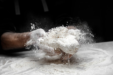 Baker chef sprinkling flour fresh dough on kitchen table, black background