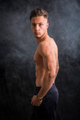 Handsome shirtless athletic young man wearing only pants, with blue eyes, looking at camera in studio shot, on dark background