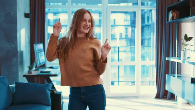 Charming caucasian young woman in beige brown sweater dancing alone in modern living room. Attractive, beautiful, cute, freedom, fun, funny girl, home, indoors. Slow motion.
