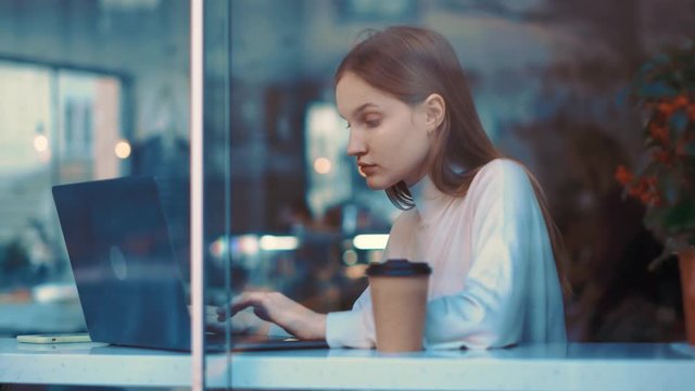 Interested pretty teenager girl in white sweater using phone in coffee shop, texing message, watcing video, contents or bloggs, scrolling, tapping. View through the window, rotation, slow motion.