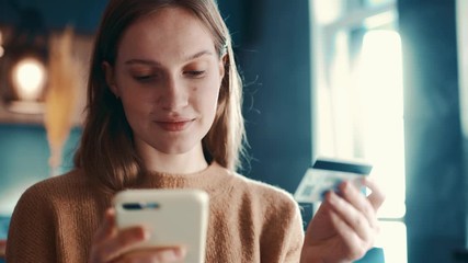 Pretty blondehair girl sitting on sofa in modern room, apartment and makes online payments through the Internet from bank card on smartphone. Online banking with smart phone. Shoot on ARRI ALEXA.