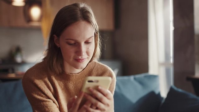 Attractive redhair teenager girl sitting of the sofa at home using phone and smiling. Feeling happy. Communication female looking message at cell phone or smartphone. Slow motion.