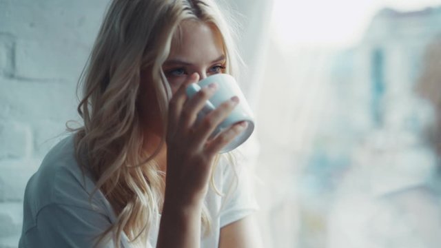 Happy beautiful woman smiling and drinking a cup of coffee or tea near the window relax in the morning cozy young home enjoying attractive comfortable relax warm slow motion