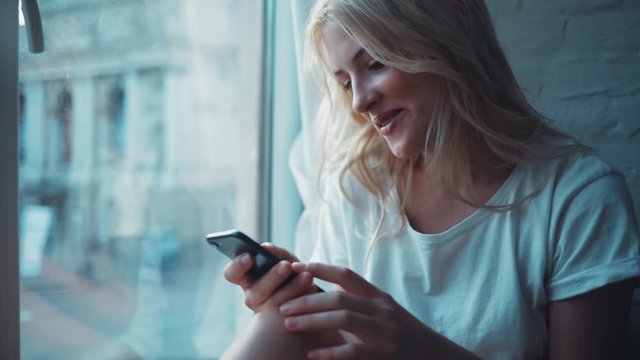Happy young woman sitting on a windowsill use phone communication female message cellphone cheerful smile internet modern smartphone portrait close up slow motion