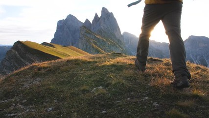 Young man hiking at Seceda mountain arriving to peak at sunrise. Backpack, yellow jacket, boots, beanie. Traveling to puez Odle, Dolomites, Trentino, Italy. - Powered by Adobe