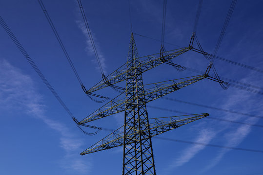 A power pole stands with many cables in front of a blue sky with few clouds when the sun is shining in Germany