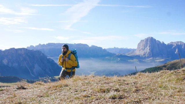 Young man hiking at Seceda mountain arriving to peak at sunrise. Backpack, yellow jacket, boots, beanie. Traveling to puez Odle, Dolomites, Trentino, Italy.