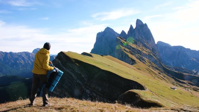 Young man hiking at Seceda mountain taking off backpack on the peak at sunrise. ellow jacket, boots, beanie. Traveling to puez Odle, Dolomites, Trentino, Italy.