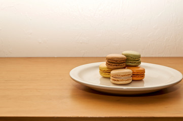 Five small colorful french macaron cookies of different colors and flavors on white plate or dish on wooden table with stuccoed wall. 