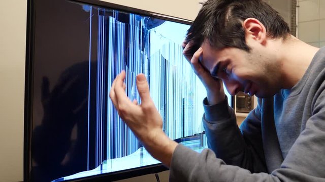 Frustrated man looking at his broken flat screen TV and trying to fix it without success.