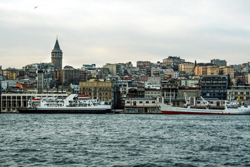 Galata Tower, on its hill, in Karakoy and Beyoglu district, taken during a cloudy winter afternoon, while the sea, ferry boats and cargo ships can be visible in foreground in Istanbul, Turkey