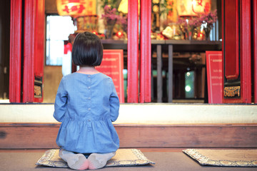 Back portrait little girl sits on the floor and prays in front of Chinese temple with blurred background