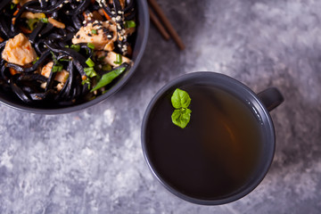 Black squid ink udon noodles with chicken meat in bowl with chopsticks and cup of mint tea on dark stone background. Top view.