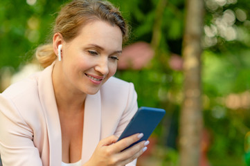 Fototapeta premium Close-up portrait of a smiling woman with smartphone on the street. Happy businesswoman is using phone, outdoors. Cheerful businesswoman in a jacket with cell phone in park.