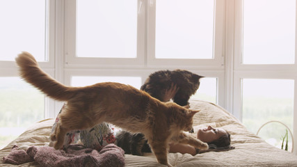 Young pretty woman relaxing with her lovely Maine Coon cat laying in bed by the window