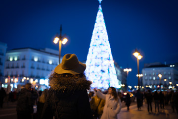 Obraz premium Tourist woman with warm clothes looking at Puerta del Sol square illuminated by christmas lights and a shinny christmas tree.
