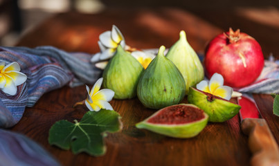 Naklejka na ściany i meble Fresh whole and cut fruits figs, pomegranate and exotic plumeria flowers on wooden table background. Still life