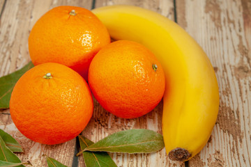 Three ripe tangerines and banana on a wooden background next to green leaves close-up with selective focus.