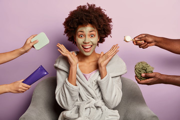 Shot of dark skinned female model has flawless skin, applies clay mask on face, spreads palms over head, wears comfortable robe, surrounded with beauty products, gets ready for date with boyfriend