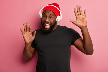Lets enjoy out party. Glad bearded Afro American man uses modern wireless headphones, listens...
