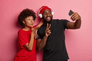 Lovely Afro woman sends air kiss, man makes peace gesture, pose for making selfie, look gladfully at smartphone device, stand together in studio over pink background, listen music in headset
