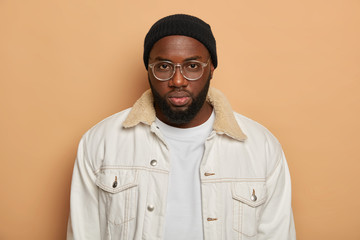 Studio shot of handsome dark skinned man with thick bristle wears transparent glasses, white shirt, black hat looks seriously at camera, being hard to impress, has calm expression, poses on beige wall