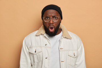 Shocked emotional bug eyed man stares at camera with omg expression, expresses astonishment and great wonder, wears big spectacles, white clothes, isolated over beige background. Reaction, emotions