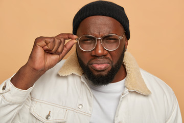 Headshot of unshaven sad black man looks through glasses scrupulously, tries to see something, has...