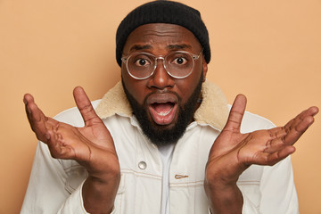 Close up shot of puzzled bearded man full of doubts, spreads palms and looks with bewilderment,...
