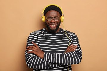 Glad dark skinned man laughs happily, keeps arms folded, wears striped jumper and transparent glasses, stereo yellow headphones, being music fan, enjoys favourite hobby, isolated on beige wall