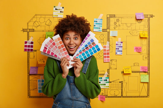 Joyful dark skinned curly woman chooses color for painting room in new house, looks through palette samples, stands against home sketch, has pleasant smile. Repair, building and renovation concept