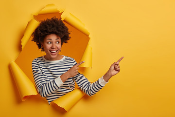 Studio shot of attractive curly girl шn striped jumper points aside on blank space with both index fingers, stands in paper hole of yellow background, happy to show cool promotion or advert.