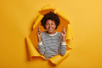 Hopeful woman with Afro hairstyle keeps fingers crossed, prays for good fortune, smiles happily,...