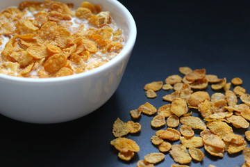 Corn flakes with milk in a white round bowl on a black metallic surface. Copy space for text.