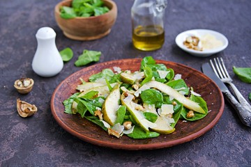 Green salad of fresh spinach, juicy pear, walnuts and parmesan on a brown clay plate on a dark concrete background. Vegan recipes. Healthly food. Selective focus