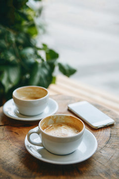 Two cups of coffee and a cellphone on a wooden table in a cozy cafe. Concept of meeting people or morning coffee drinking.