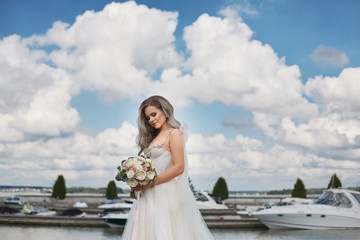 Fototapeta na wymiar Beautiful young bride with platinum blond hair in lace wedding dress keeps bouquet of fresh flowers and posing on the lake coast. Young woman with wedding hairstyle in stylish dress posing outdoors.
