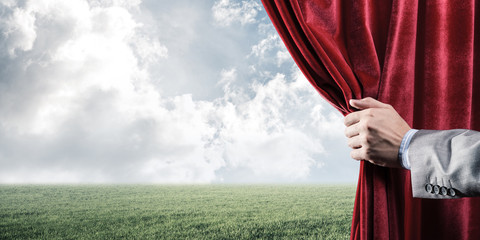 Fototapeta premium Green meadow behind red curtain and hand holding it