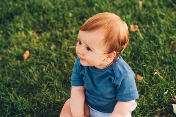 Outdoor portrait of adorable redheaded baby boy playing in summer parc, sitting on green grass, wearing blue bodysuit