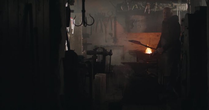 Blacksmith hammering heated iron on anvil making  knife dark eerie haze