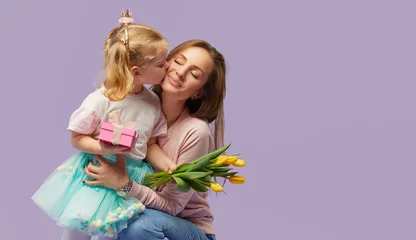 8 march card! Little daughter kisses and hugging her mother with yellow flowers tulip and gift box. Purple studio background. Mothers day concept © oes