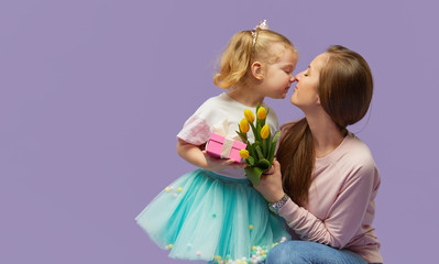 8 march card! Little daughter kisses and hugging her mother with yellow flowers tulip and gift box. Purple studio background. Mothers day concept