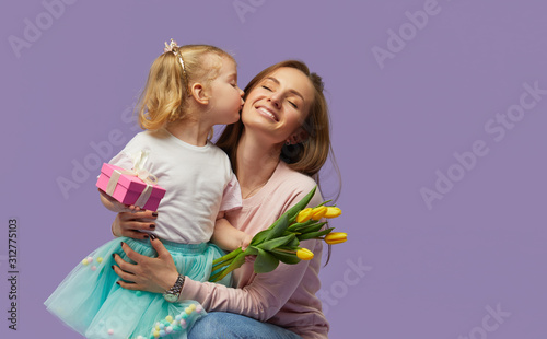 8 march card! Little daughter kisses and hugging her mother with yellow flowers tulip and gift box. Purple studio background. Mothers day concept