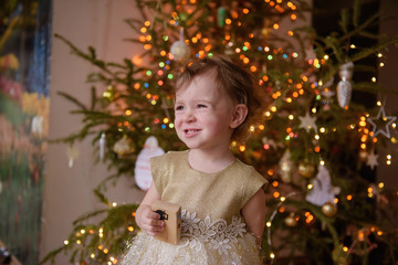 Beautiful little blonde girl smiles at the Christmas tree on Christmas Eve in a gold sequin dress. The concept of celebrating the new year at home. Waiting for a miracle and Santa Claus. Hand made toy