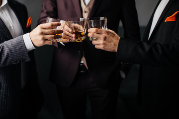 Close-up partial view of three friends clink glasses of whiskey drink, alcoholic beverage. Men in formal wear toasting with glasses of whiskey. Whisky drink