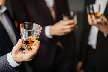 Close-up partial view of three friends clink glasses of whiskey drink, alcoholic beverage. Men in formal wear toasting with glasses of whiskey. Whisky drink