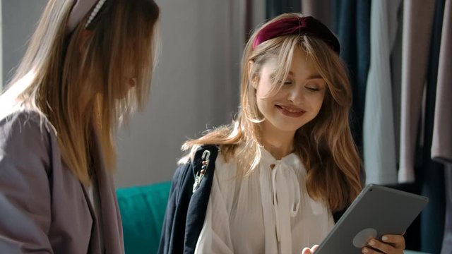 Happy young women looking at each other and nodding while choosing garments in online catalog while sitting on couch in store online.