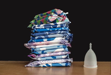 A silicone menstrual cup next to a stack of sanitary towels and tampons.  Sustainability / zero waste / frugality concept. Alternative way to manage periods / menstrual cycles. 